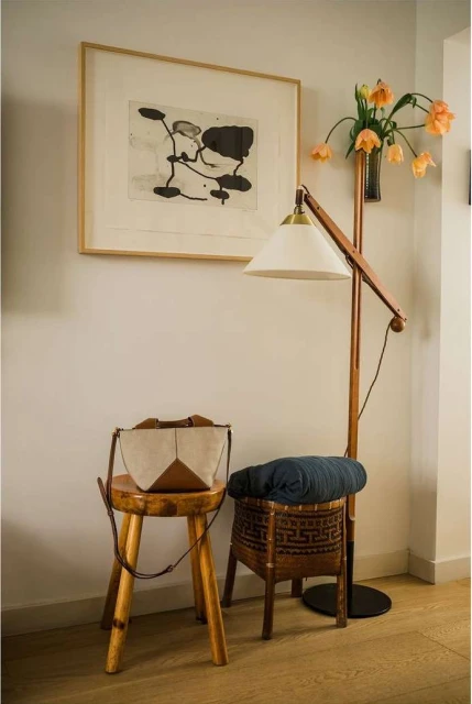 Living room corner with a wooden stool, a woven basket holding a blanket, a standing lamp, a framed art print, and flowers in a vase.