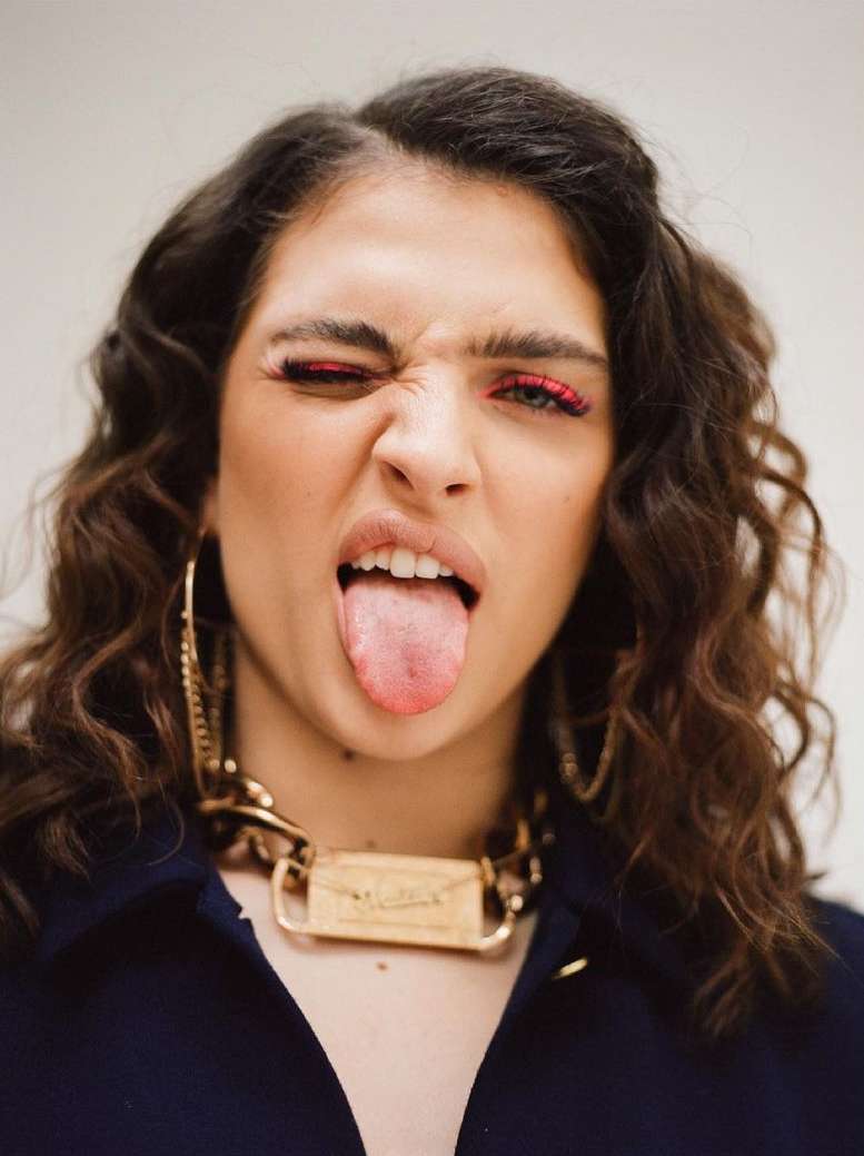 Close-up portrait of a young adult woman sticking her tongue out and winking with red eyeliner, against a neutral background.