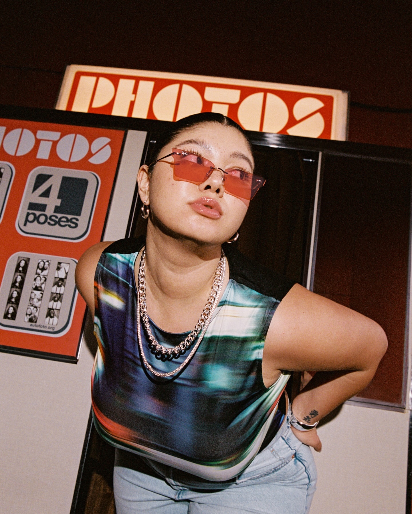 Young woman with sunglasses and layered necklaces poses in a photo booth with a retro aesthetic and eye-level angle.