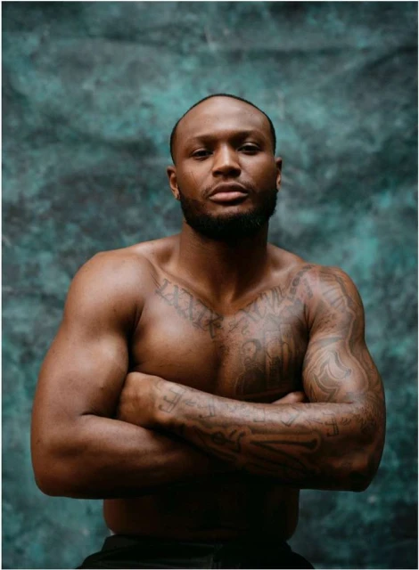 Portrait of a muscular, shirtless adult male with tattoos, arms crossed, and serious facial expression against a mottled teal background.