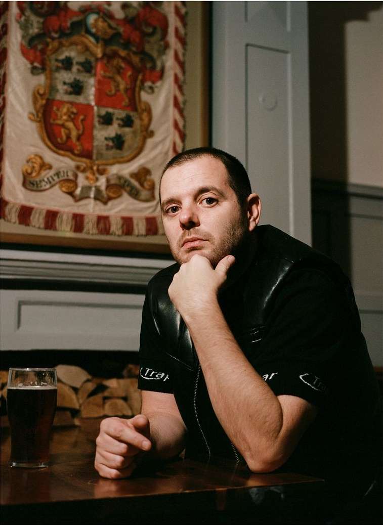 Indoor portrait of an adult male sitting at a table with a glass of dark beer, wearing black, with a banner in the background.