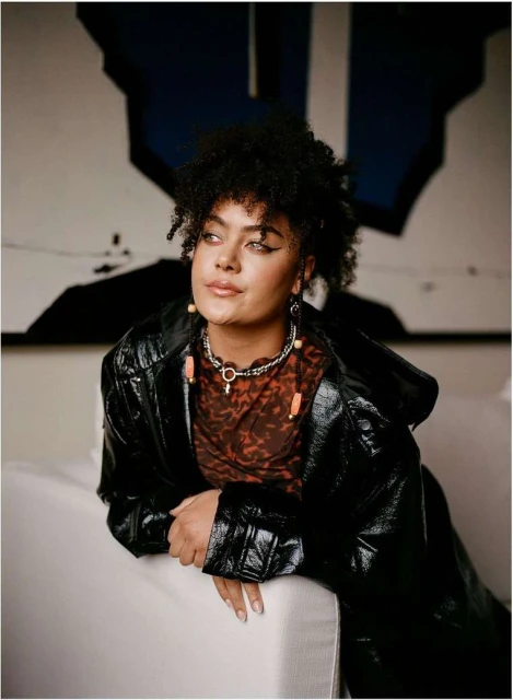 Portrait of a woman with afro hairstyle in a shiny black coat leans against a white sofa, gazing to the side with an abstract painting backdrop.