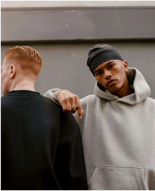 Two young adult men pose together indoors against a gray wall, one wearing a gray hoodie and the other wearing a black sweatshirt.