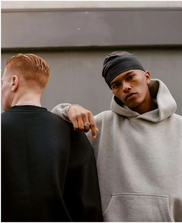 Two young adult men pose together indoors against a gray wall, one wearing a gray hoodie and the other wearing a black sweatshirt.