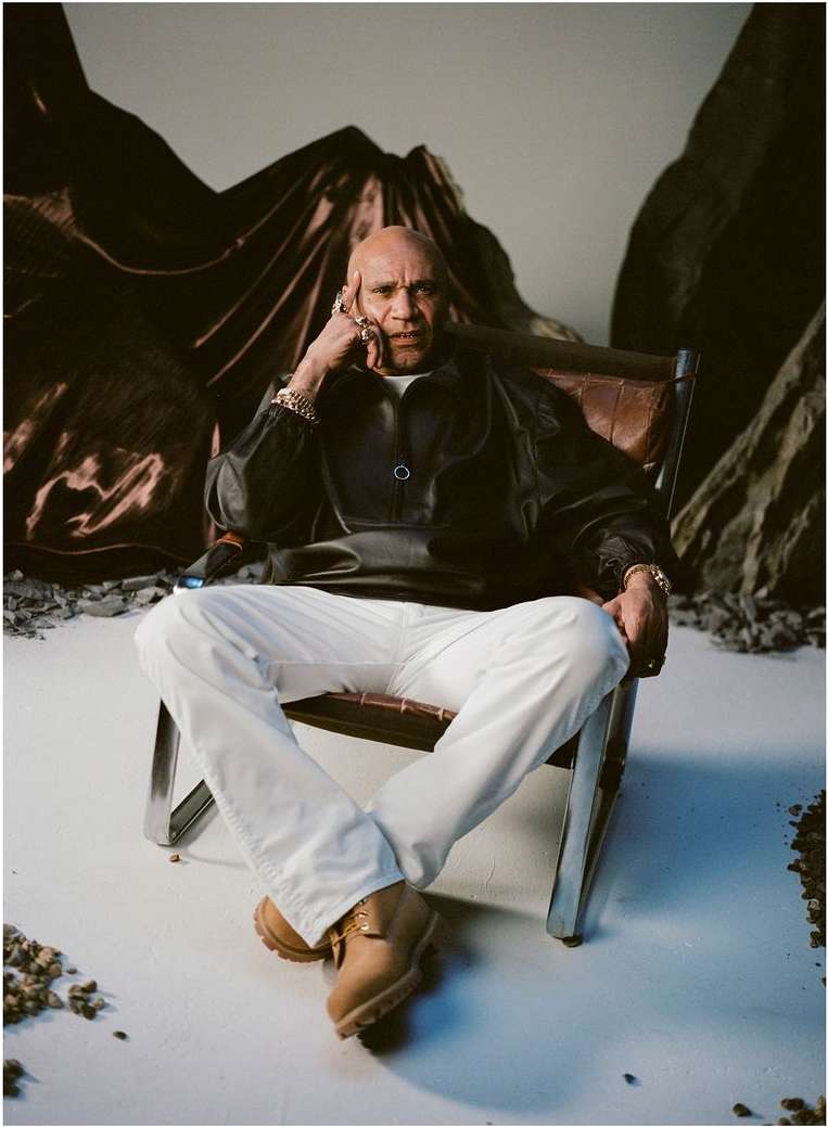 Stylish bald man in black jacket and white jeans sits in leather chair in studio with backdrop, wearing jewelry and tan work boots.