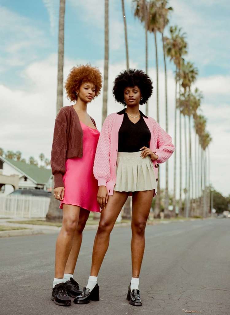 Two young women stand on a palm-lined street wearing cardigans, skirts, dresses, and black shoes with light blue sky overhead.