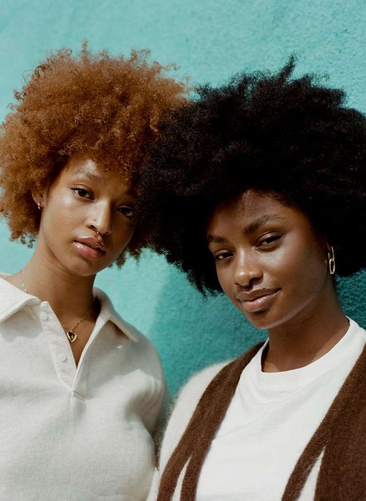Close-up portrait of two young Black women with natural hair posing in front of a turquoise wall wearing casual clothing and jewelry.