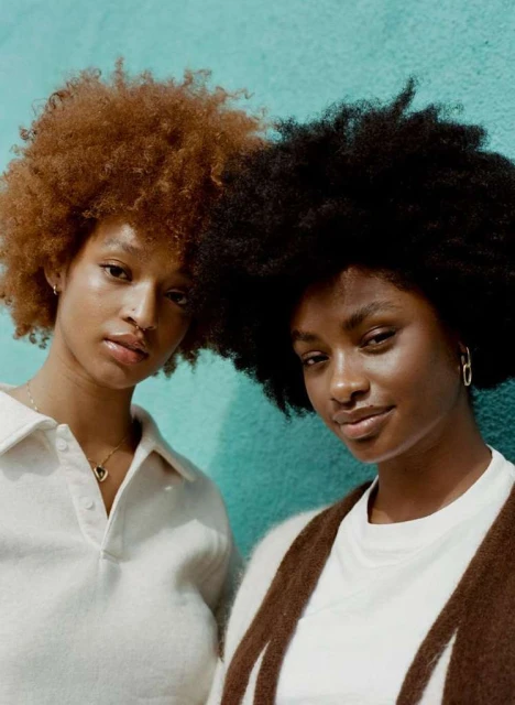 Close-up portrait of two young Black women with natural hair posing in front of a turquoise wall wearing casual clothing and jewelry.