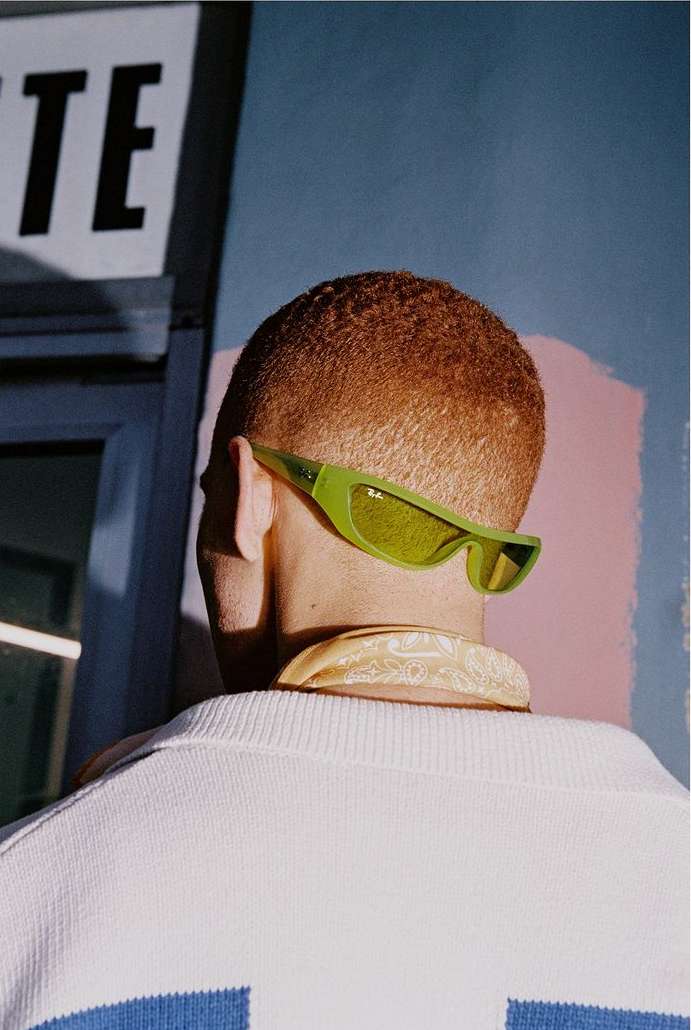 Back view of a man with a buzz cut wearing green sunglasses and a white sweater with a blue stripe, set against a blue and pink wall.