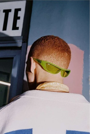 Back view of a man with a buzz cut wearing green sunglasses and a white sweater with a blue stripe, set against a blue and pink wall.