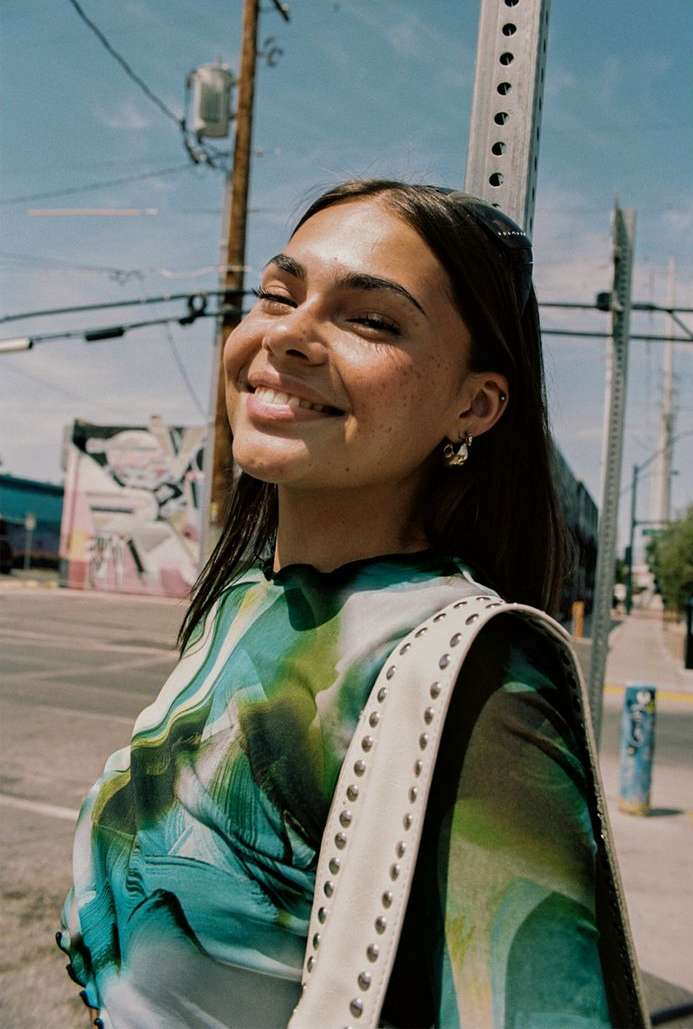 Smiling woman with fair skin and a patterned top stands outdoors on a sunny day, with a white purse and earrings.