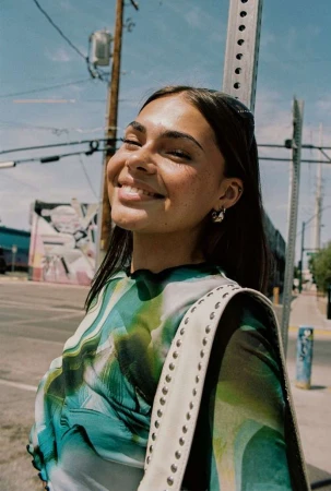 Smiling woman with fair skin and a patterned top stands outdoors on a sunny day, with a white purse and earrings.