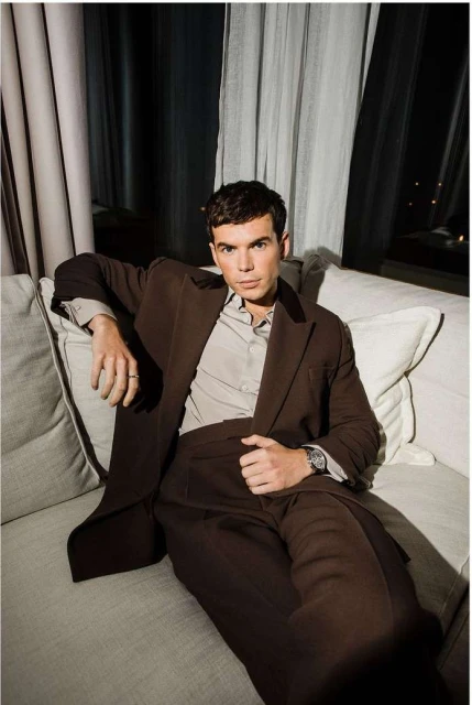 Adult male in a brown suit reclines on a gray sofa in an indoor setting with neutral curtains and natural light from a nearby window.