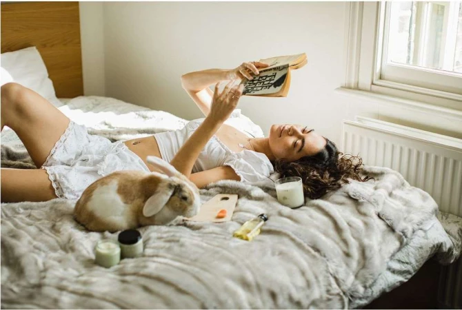 A young woman is reading a book on a bed with a rabbit, cosmetics, and a candle, bathed in soft natural light from a nearby window.