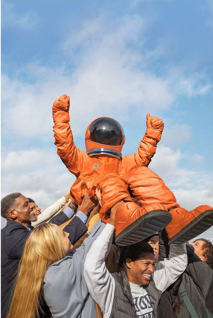 An astronaut in an orange space suit being held up by a group of adults against a blue sky with clouds, celebrating, triumphant.