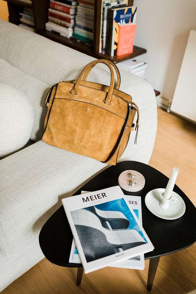 Tan suede tote bag sitting on a light grey couch next to a black side table with books and a candle, indoor setting.