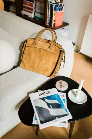 Tan suede tote bag sitting on a light grey couch next to a black side table with books and a candle, indoor setting.
