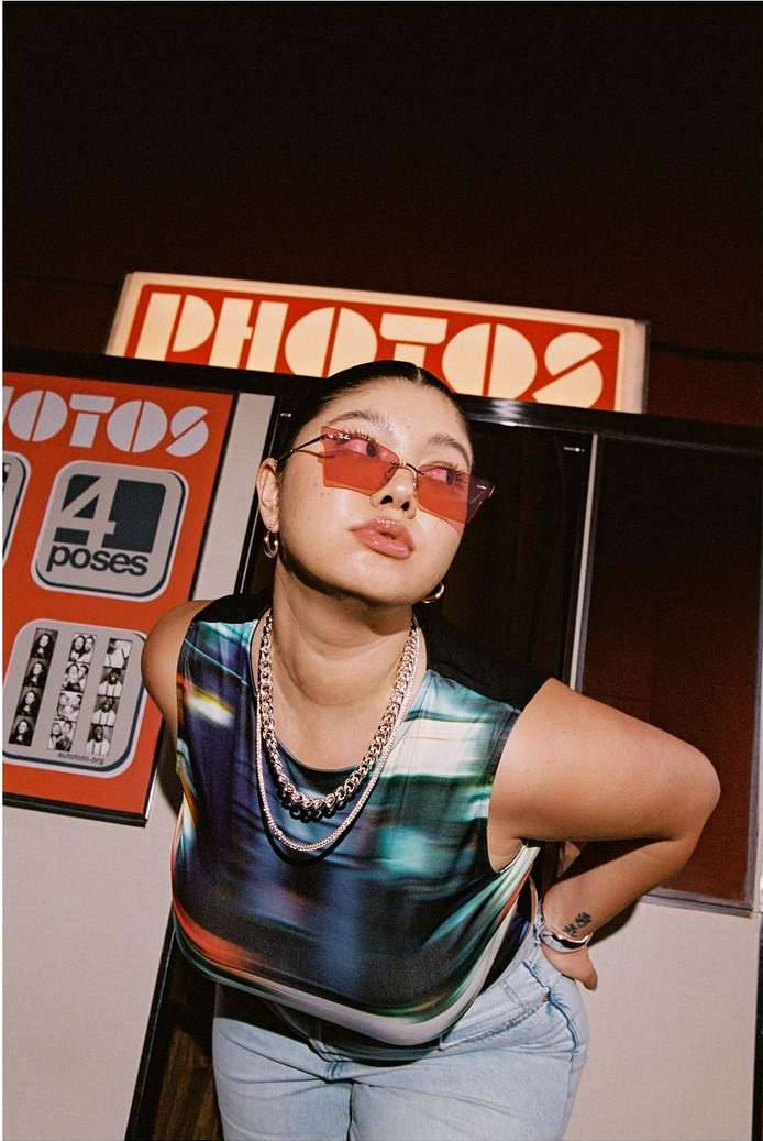 Woman poses in front of a vintage photo booth with red sunglasses, a colorful top, jeans and layered silver necklaces in a studio.