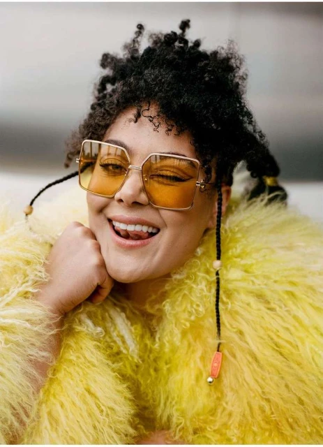 Close-up of a smiling woman with curly hair, wearing yellow-tinted glasses and a fluffy yellow jacket, against a neutral backdrop.