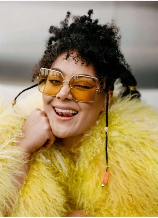 Close-up of a smiling woman with curly hair, wearing yellow-tinted glasses and a fluffy yellow jacket, against a neutral backdrop.