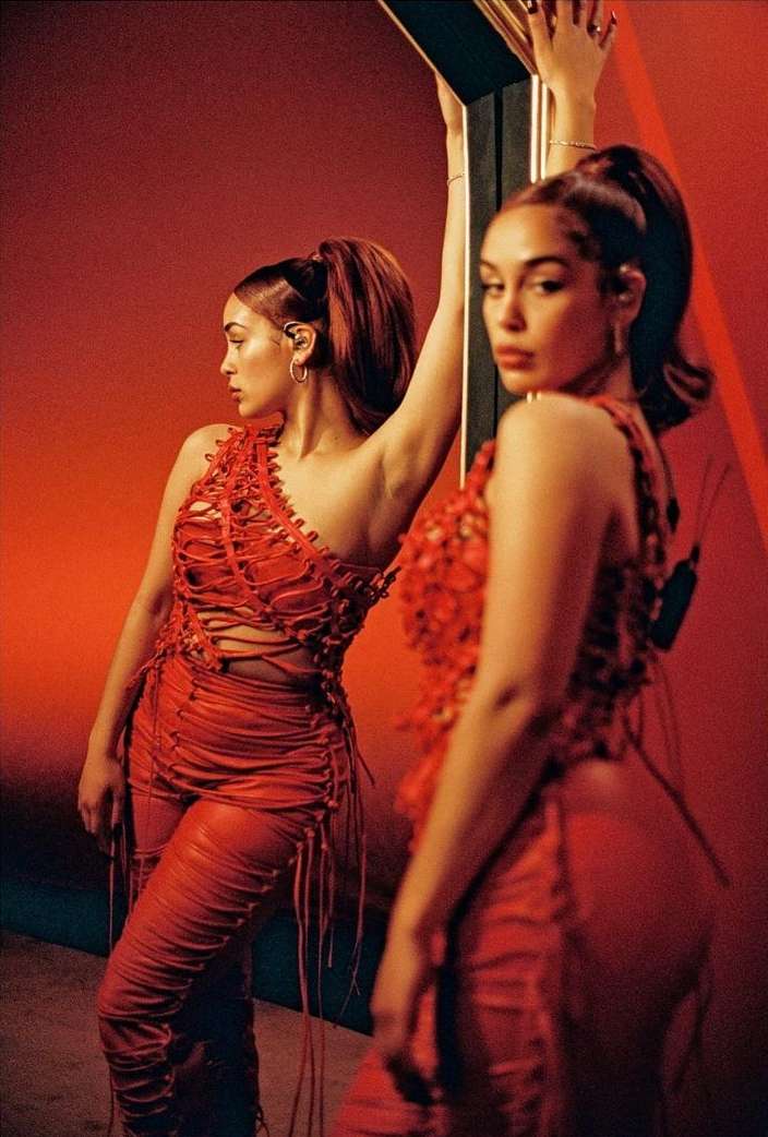 An adult woman leans against a mirror reflecting her, dressed in a red outfit with a high ponytail against a matching red background.
