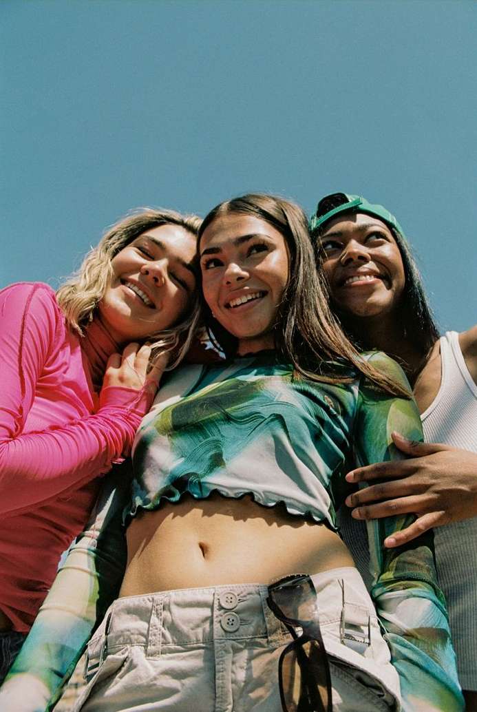 Low angle shot of three young women of diverse ethnicities smiling outdoors with blue sky background, wearing casual tops and accessories.