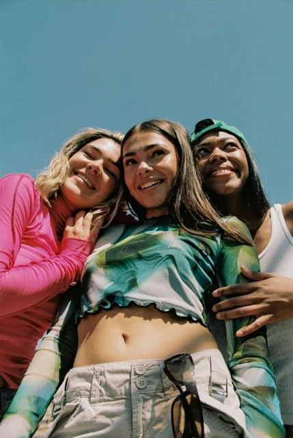 Low angle shot of three young women of diverse ethnicities smiling outdoors with blue sky background, wearing casual tops and accessories.
