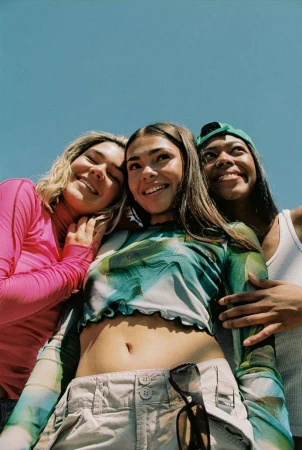 Low angle shot of three young women of diverse ethnicities smiling outdoors with blue sky background, wearing casual tops and accessories.