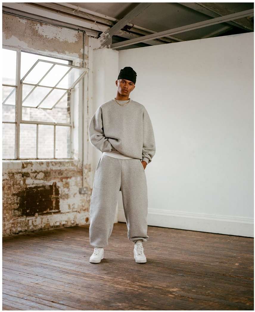 Young adult in a gray sweat suit and black beanie stands in a minimalist interior with wooden floors and a large window.