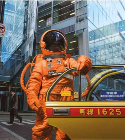 An astronaut in an orange spacesuit stands next to a yellow taxi on a city street with modern office buildings in the background.