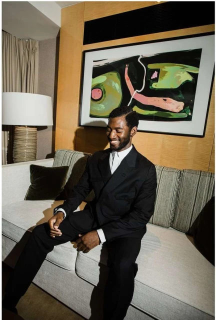 Portrait of an adult man in a dark suit sitting on a couch with a smile, featuring a modern art painting and lamp in the background.