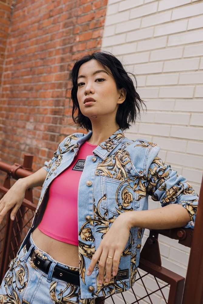 Young Asian woman in fashionable denim jacket and pants posing against brick wall. Street style fashion shoot, eye-level angle.