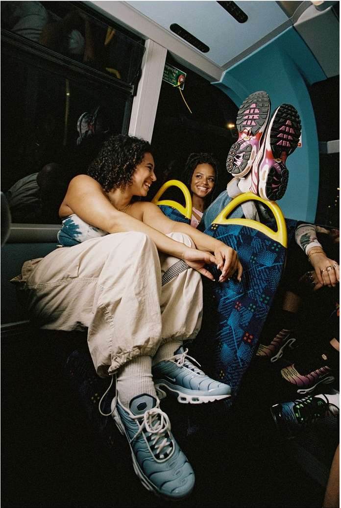 Two women sitting on a bus, one with feet propped up, wearing sneakers. Interior shot shows the bus seats and dim lighting.
