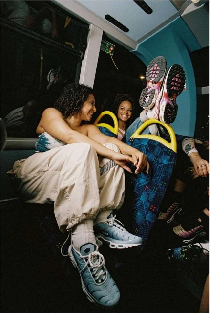 Two women sitting on a bus, one with feet propped up, wearing sneakers. Interior shot shows the bus seats and dim lighting.
