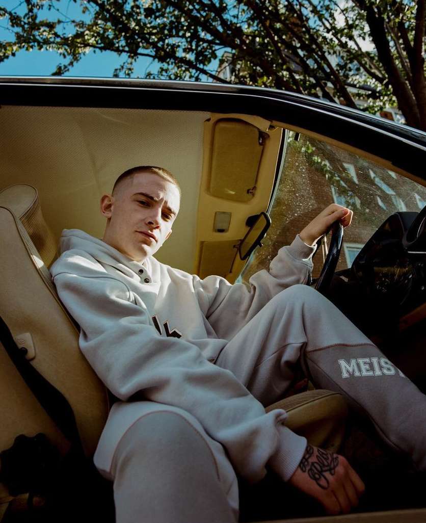 A young man in a grey sweatsuit sits in a vintage car with an open door, shot with outdoor lighting and an eye-level angle.