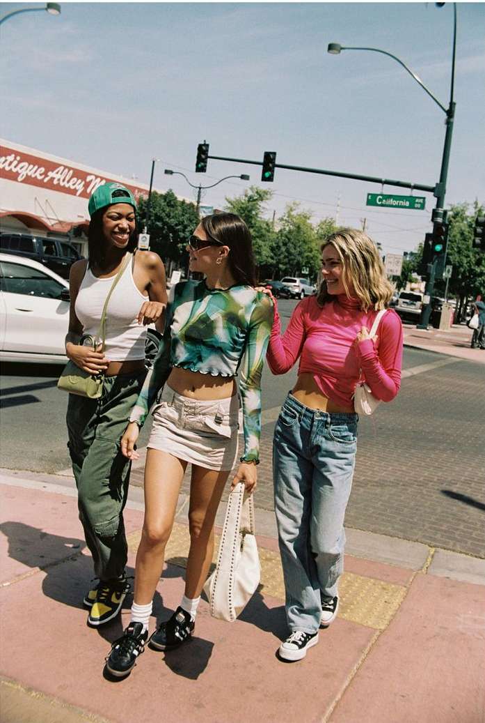 Three young women walk together on a city street with vintage clothing and bags, captured in bright daylight with a background of buildings.