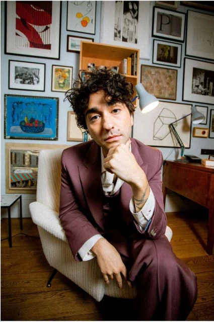 Portrait of a curly-haired man in a suit seated in a chair in a room with many framed pictures on the wall behind him.