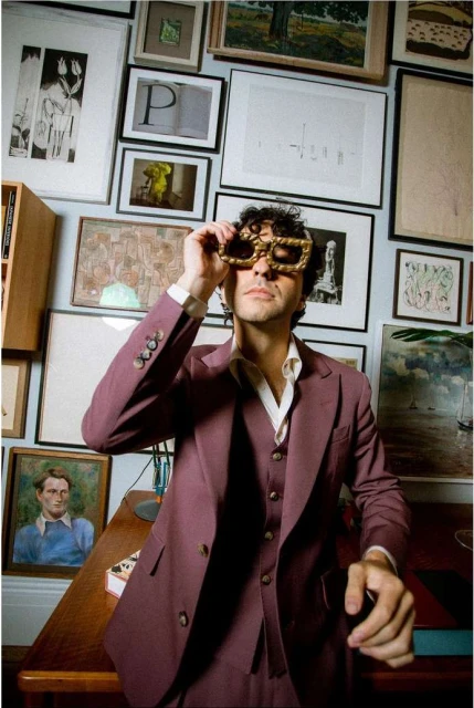A man in a maroon suit and oversized gold glasses poses in front of a wall of framed pictures in an indoor setting with warm lighting.