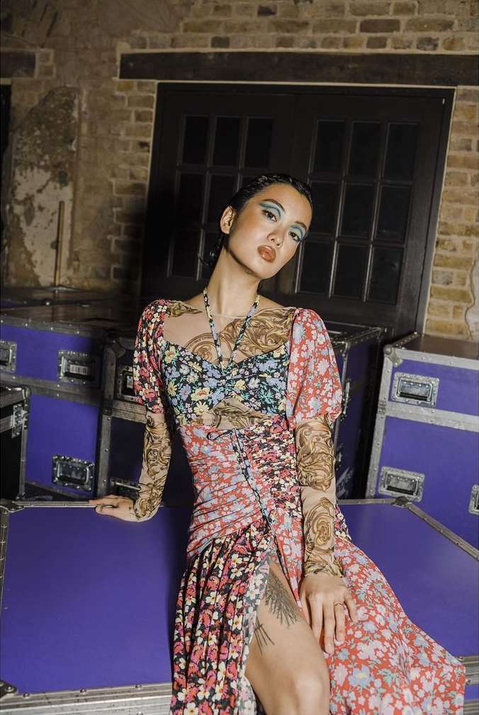 A woman in a floral-print dress poses against a backdrop of stacked cases with brick walls and a dark doorway behind her.