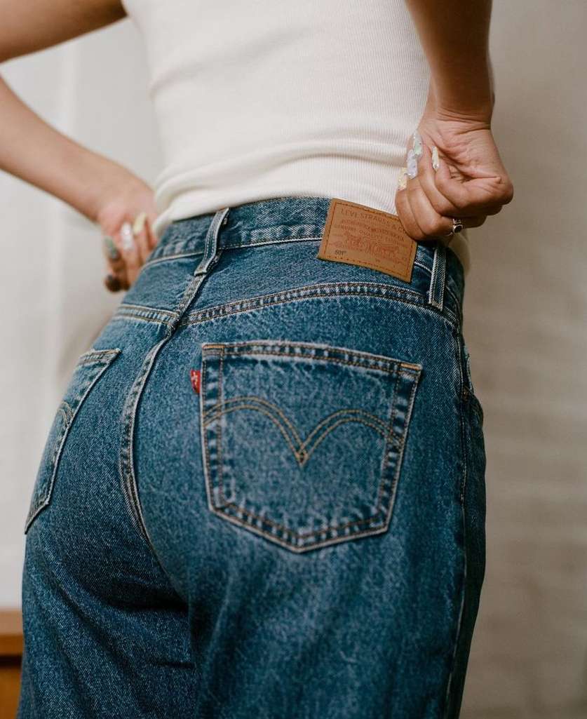 Back view of a woman in a white tank top and blue jeans, showing the classic Levi's brand tag and back pocket stitching.