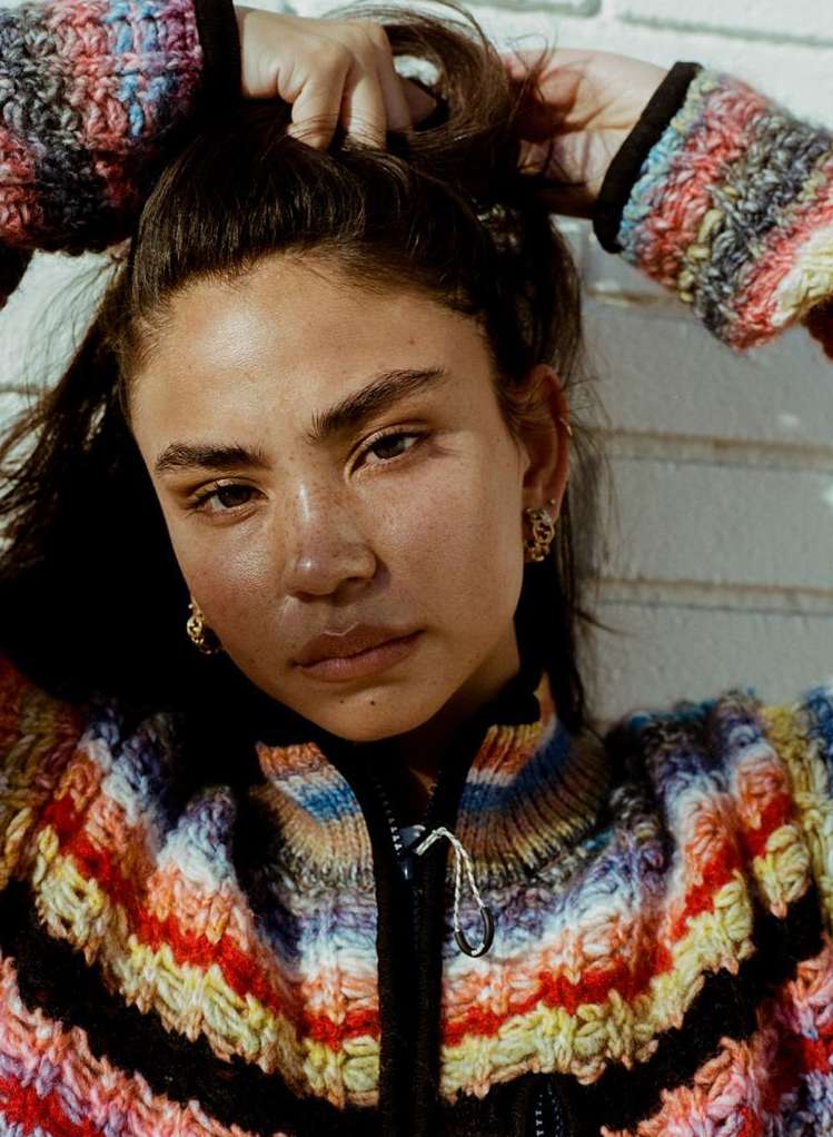 Woman with brown hair and striped knit sweater poses in a close-up shot, with a neutral facial expression against a brick wall backdrop.