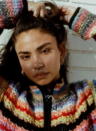 Woman with brown hair and striped knit sweater poses in a close-up shot, with a neutral facial expression against a brick wall backdrop.