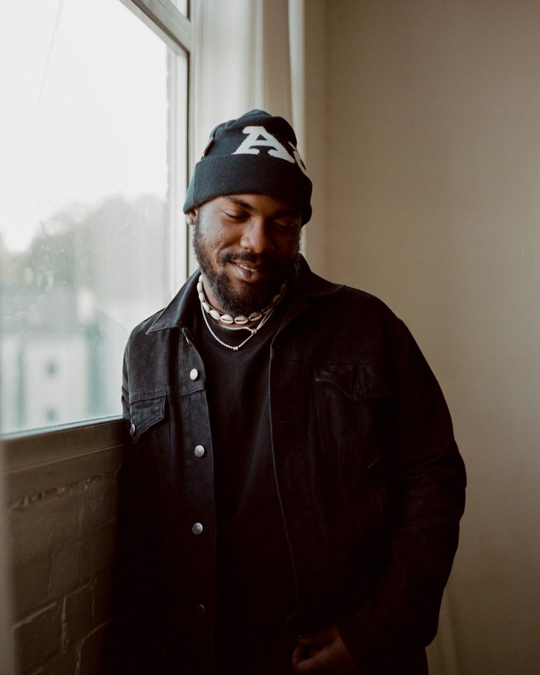 Portrait of an African American man in a black denim jacket and beanie, standing indoors near a window with natural light, looking downward.