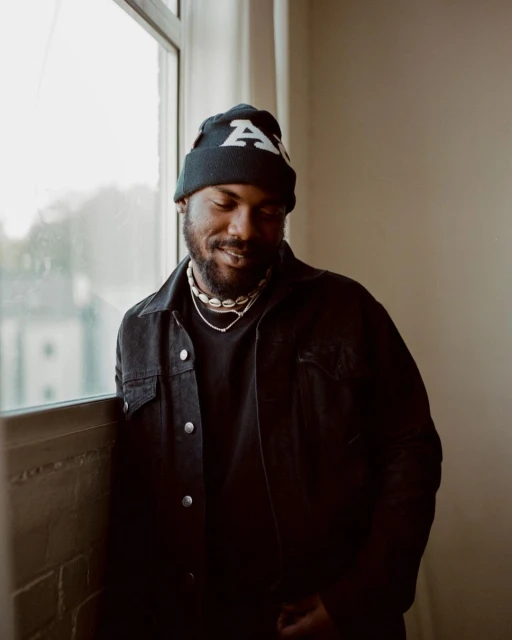 Portrait of an African American man in a black denim jacket and beanie, standing indoors near a window with natural light, looking downward.