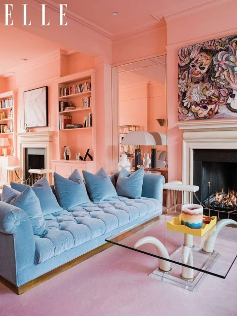 Living room interior with light blue tufted sofa, glass coffee table, fireplace, bookshelves, artwork, and pink walls and carpet.