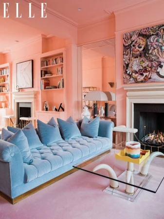 Living room interior with light blue tufted sofa, glass coffee table, fireplace, bookshelves, artwork, and pink walls and carpet.