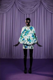 Model wearing a white and blue floral dress with black latex mask, gloves, and over-the-knee boots standing before purple drapes.