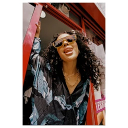 Woman with curly dark hair wearing sunglasses and patterned jacket leaning in red doorway by Vicky Grout