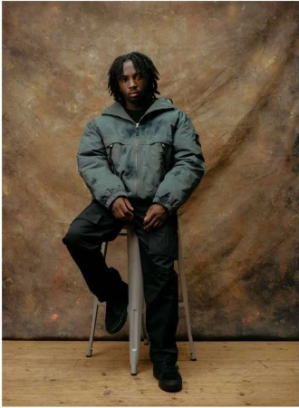Black man seated on a stool wearing a dark puffer jacket and black cargo pants against a mottled textured backdrop.
