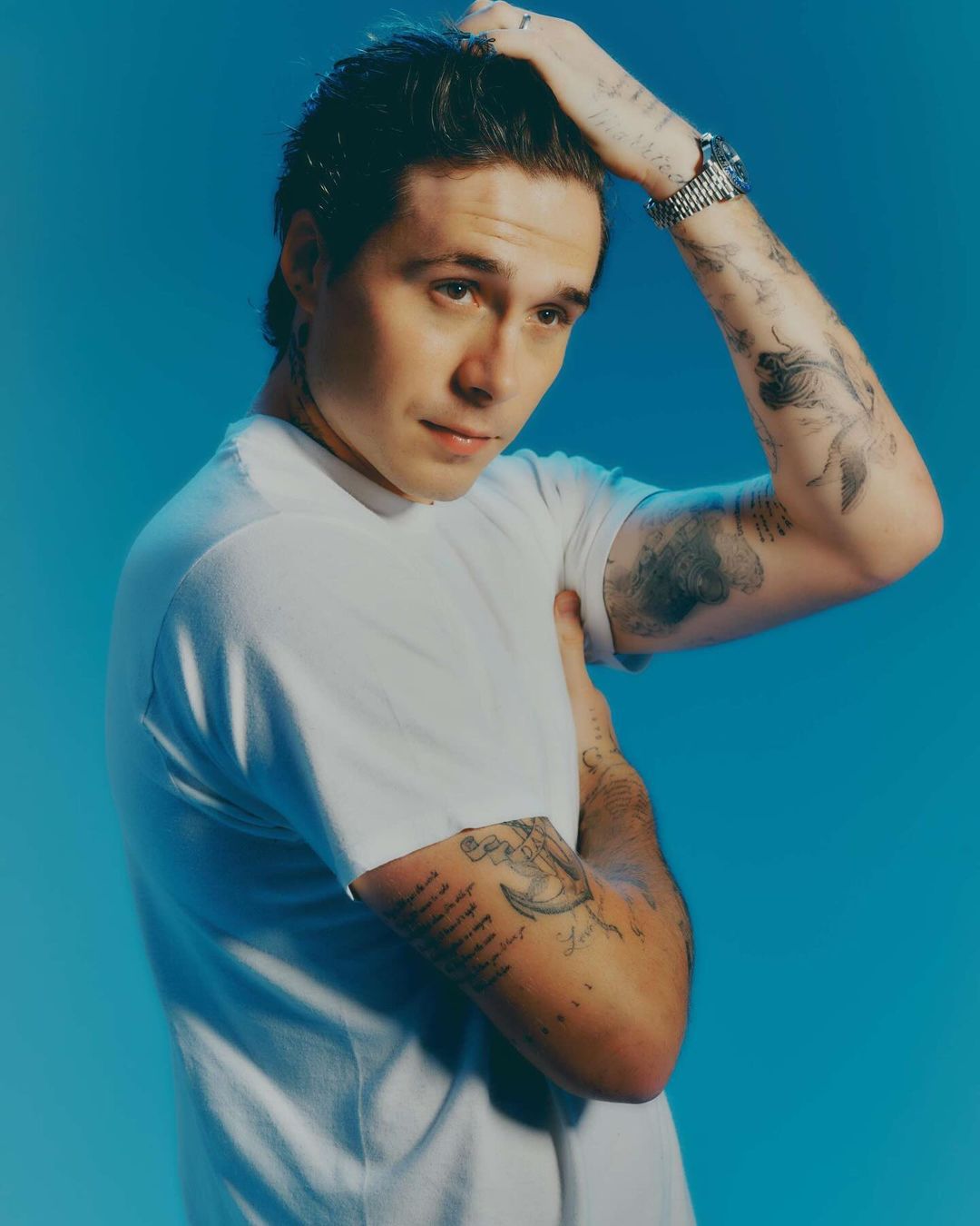 Portrait of a young adult male with dark hair in a white t-shirt posing with his hand on his head in a photo studio setting with blue background.